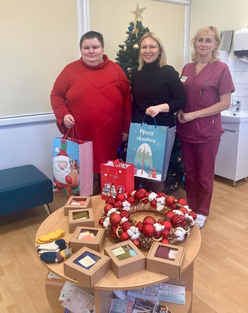 Kolm naeratavat naist seisavad jõulukuuse ees, hoides kingikotte. Esiplaanil laual on jõulupärg ja käsitöötooted, sealhulgas karbikesed ja soojad villased sokid