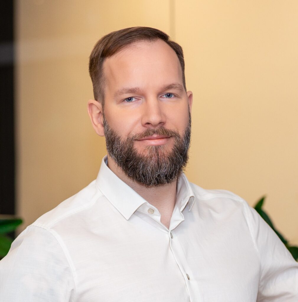 Pildil heleda särgi ja habemega noor mees kaamerasse vaatamas.