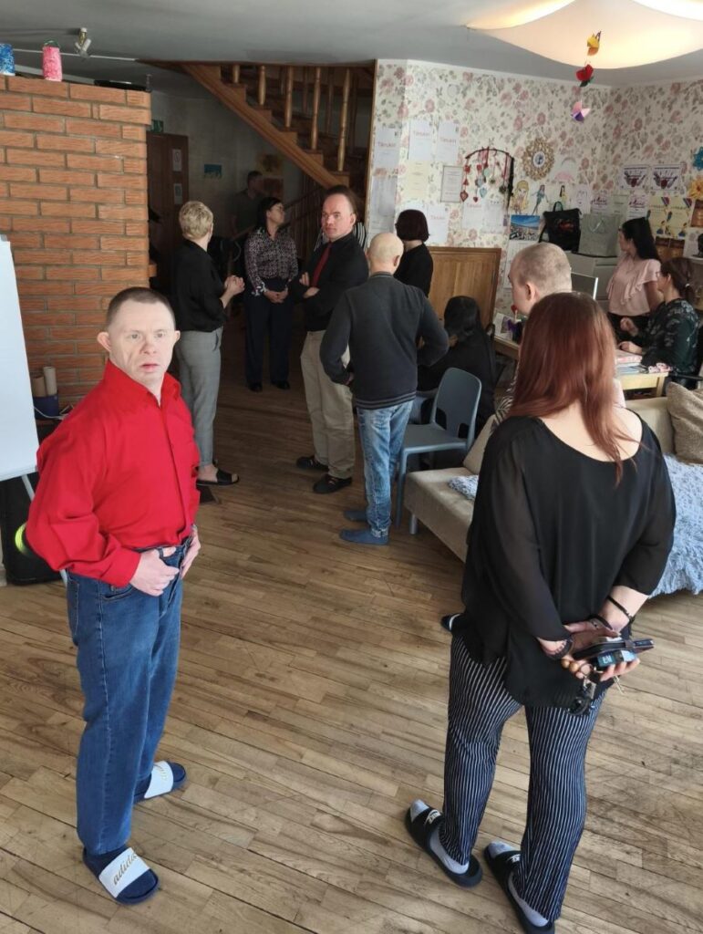 Inimesed kogunevad ja suhtlevad toas, mille seintel on joonistused ja tänukirjad. Esiplaanil seisab punases särgis mees.