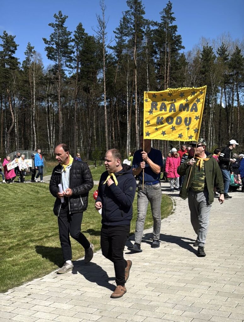 Rääma Kodu esindajad kõnnivad rõõmsalt rongkäigus kevadisel päikesepaistelisel päeval, hoides käes kollast silti kirjaga "Rääma kodu".