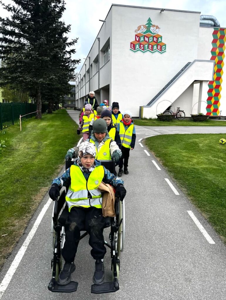 Lapsed jalutavad Vändra lasteaia juures, üks laps on ratastoolis. Seljas on helkurvestid. 