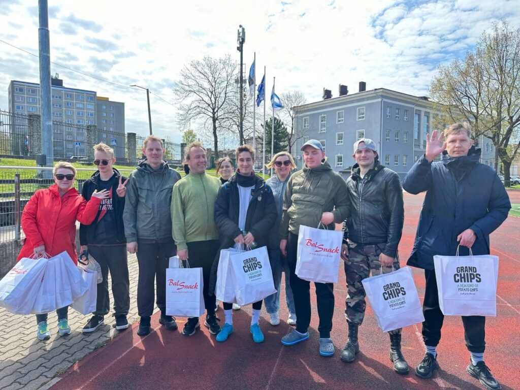 Naeratav seltskond inimesi seisab spordiväljaku ääres, käes kinkekotid kirjadega "BalSnack" ja "Grand Chips"; taustal lippude lehvides hoone ja kevadine taevas.