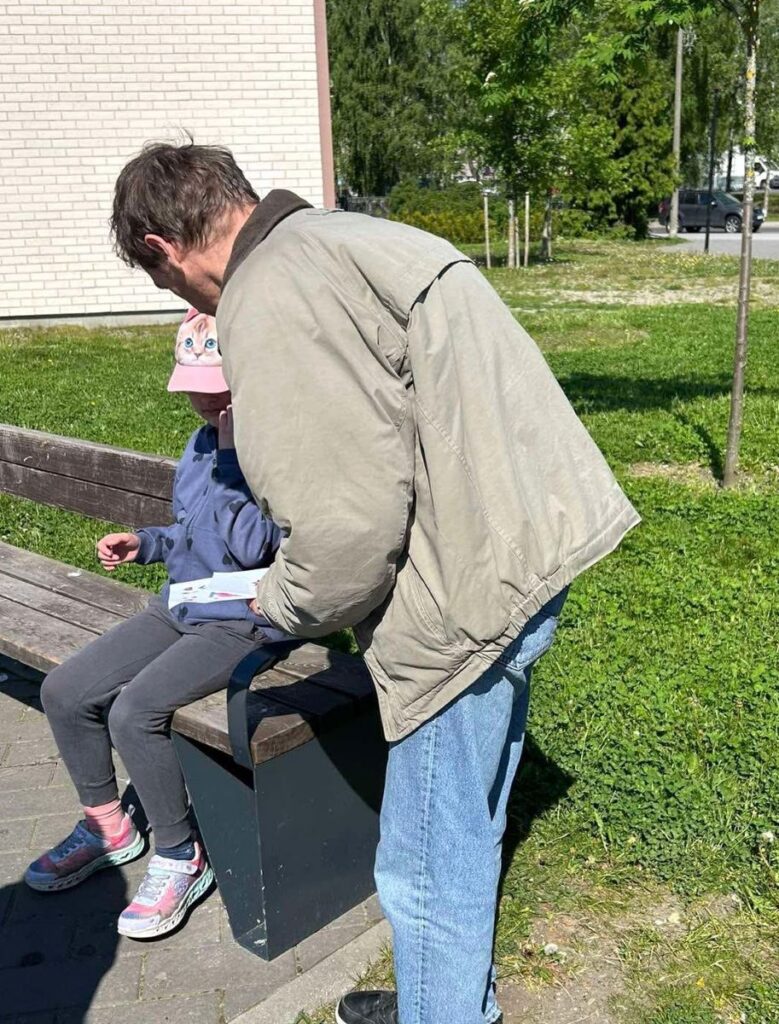Kopli Kodu elanik ulatab istuvale lapsele tervituskaardi Põhja-Tallinna lastepeol, taustal roheline muru ja päikesepaisteline päev.