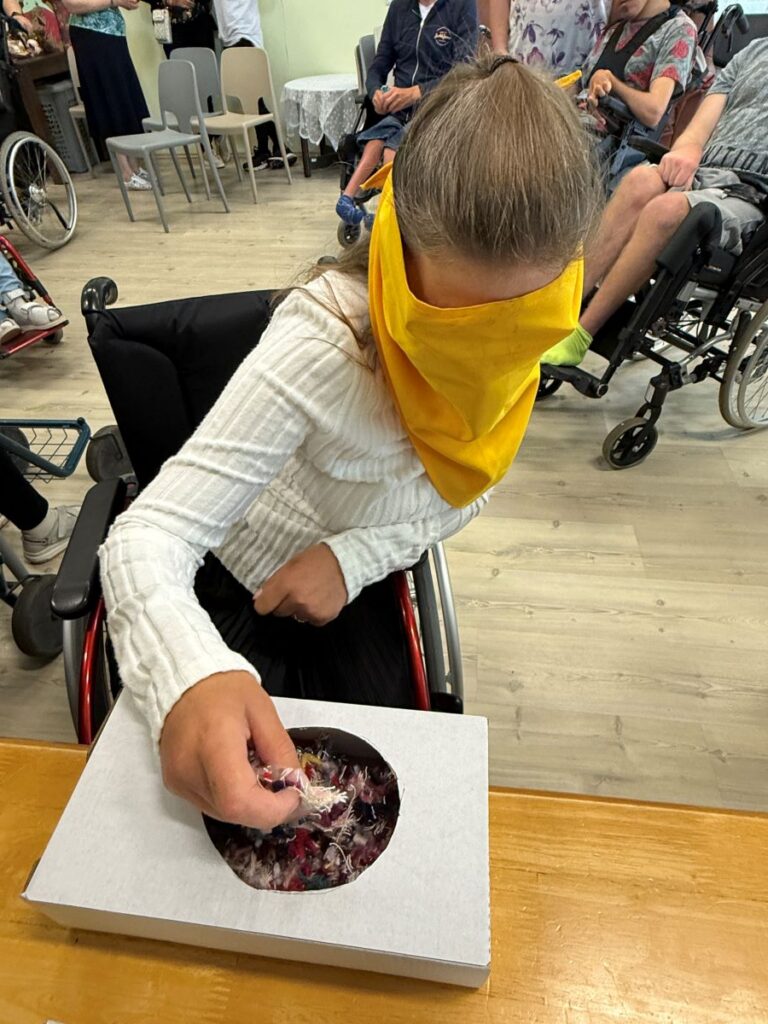 Inimesel, kes istub ratastoolis, on silmad kaetud salliga ning katsub karbis olevaid asju.
