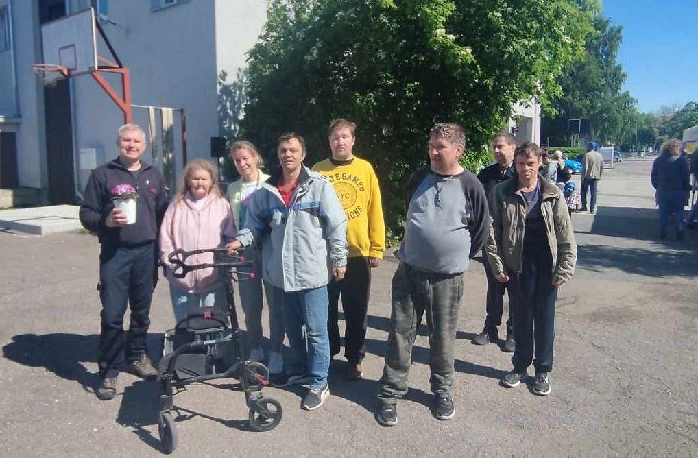 Grupipilt päikeselisel päeval: Kopli kodu elanikud koos vabatahtliku päästjaga õues, taustal päästekomando hoone ja korvpallilaud.