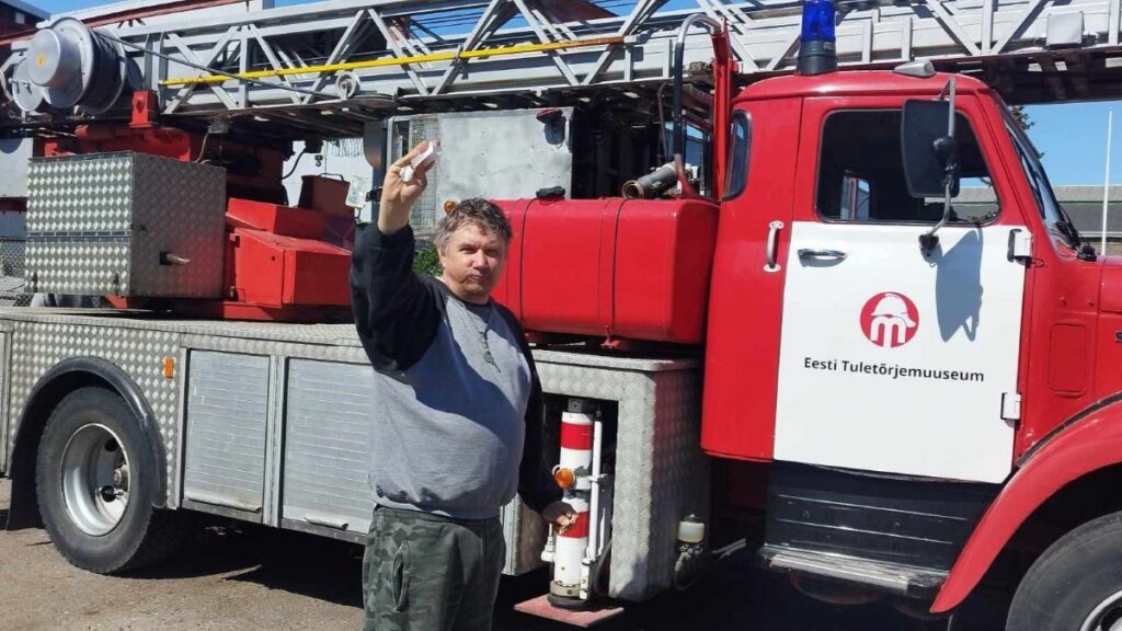 Kopli kodu elanik poseerib punase päästeauto ees, tõstes käe tervituseks.