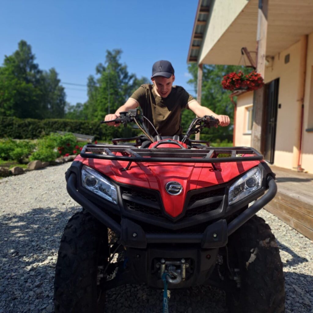 Noormees istub punasel ATV-l ja naeratab, taustal aed ja hoone