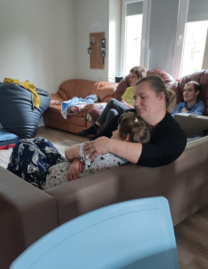  Naine hoiab teist täiskasvanut hellalt embuses, ümberringi istuvad inimesed diivanitel rahulikus ja koduses õhkkonnas