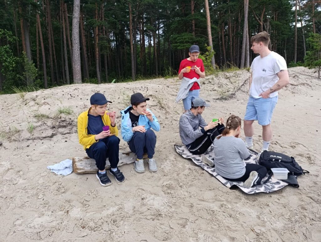 Inimesed istuvad rannaliival ja piknikutekil, söövad ja joovad männimetsa taustal.