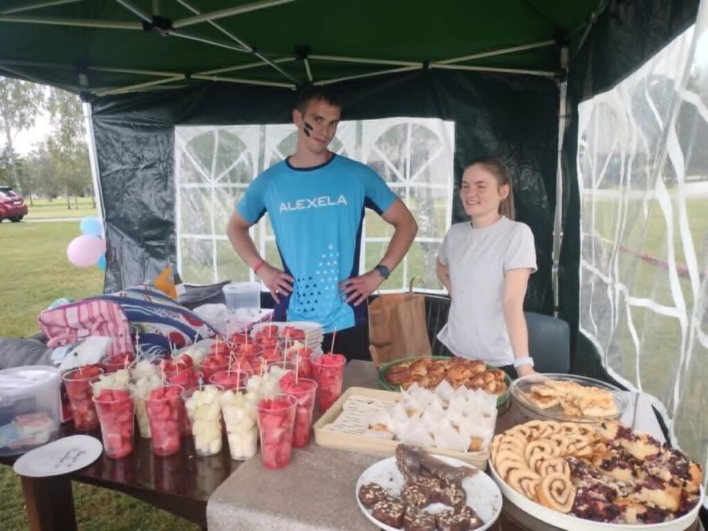 Kaks noort inimest seisavad rõõmsalt telgi all magusalauda valvamas. Laual on klaastopsid puuviljadega ning erinevad kodused küpsetised.