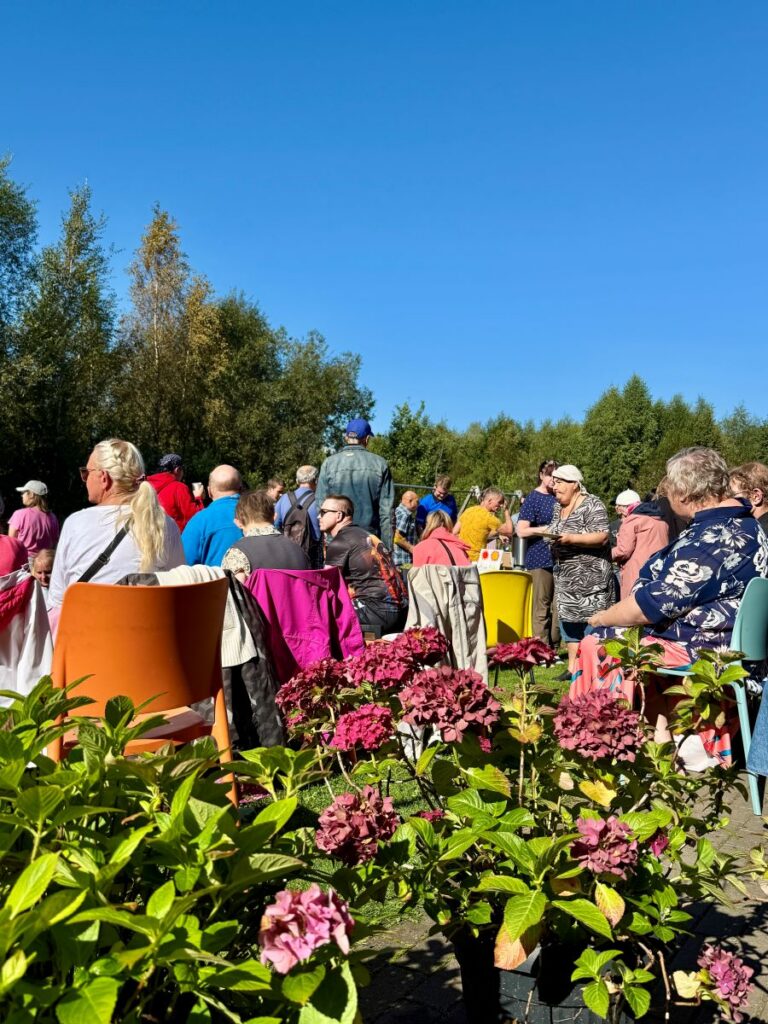 Publik istub lillede vahel murul ja jälgib rõõmsameelselt koosolemise päeva esinemist päikesepaistelisel päeval.