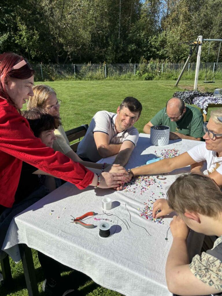 Inimesed istuvad õues laua ümber ja meisterdavad käevõrusid – laual helmed, tangid ja niidid. Nad hoiavad käsi üksteise peal kuhjas.