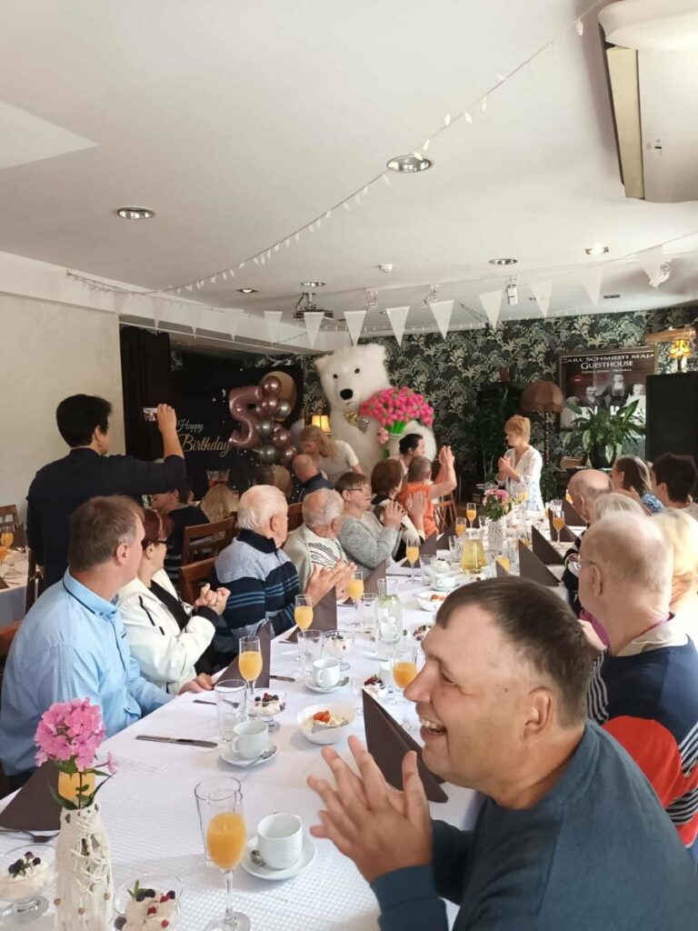 Sünnipäevalaua taga istuvad Põltsamaa Kingu kodu elanikud, taustal astub sisse suur karu lilledega.