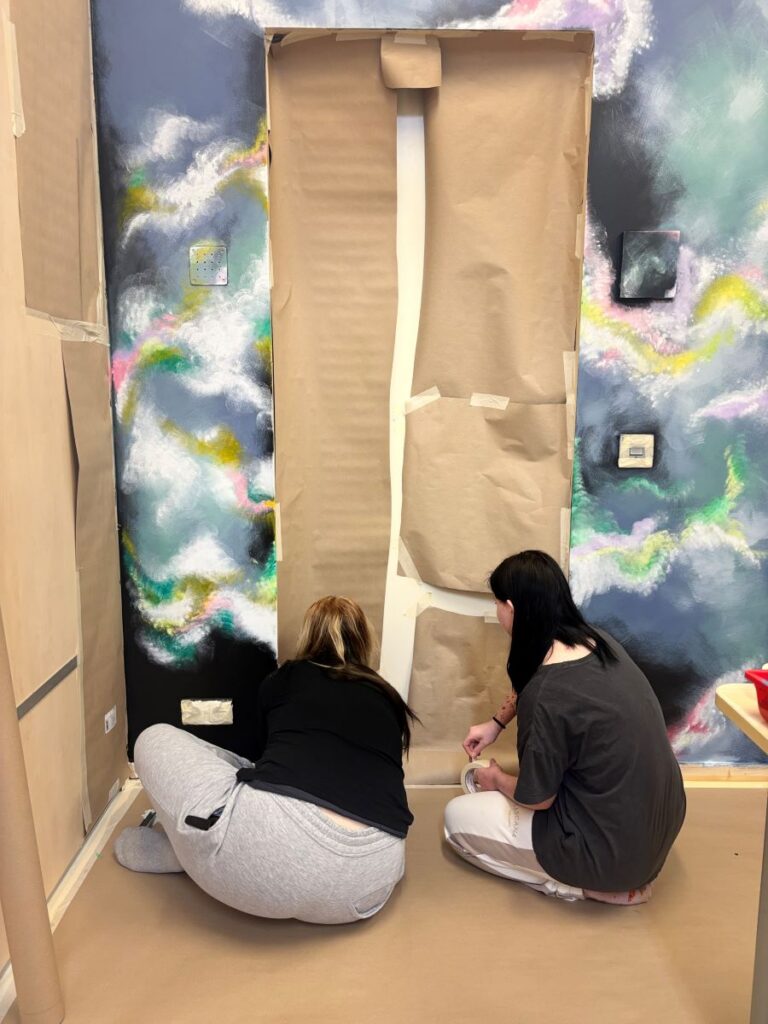 Kaks noort istuvad põrandal ja teibivad galaktika seinamaali ette pindu kinni.