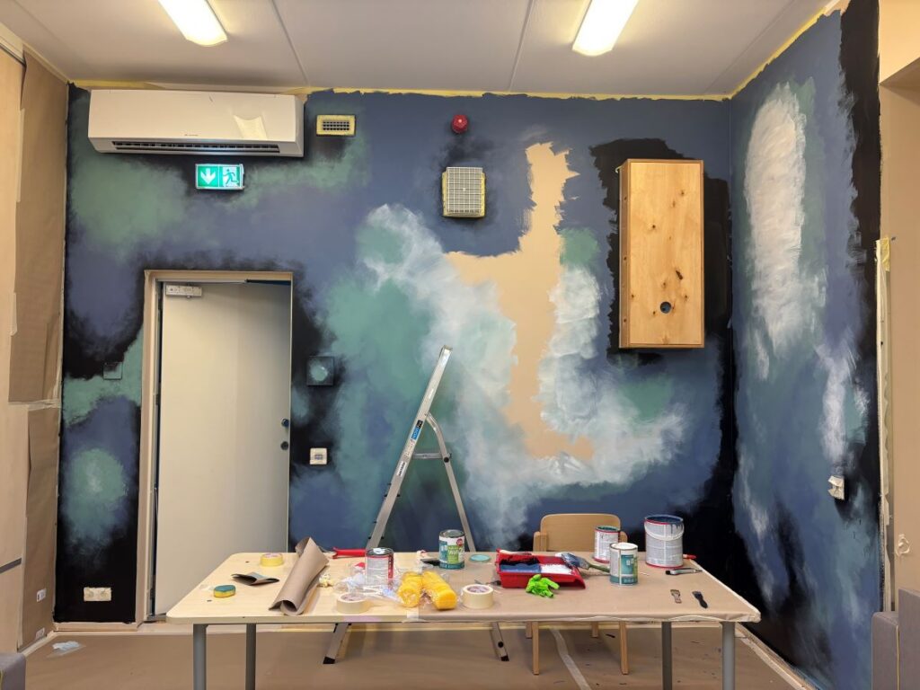 Galaktika seinamaal tööprotsessi keskel, laud ees värvipurkide ja tööriistadega.