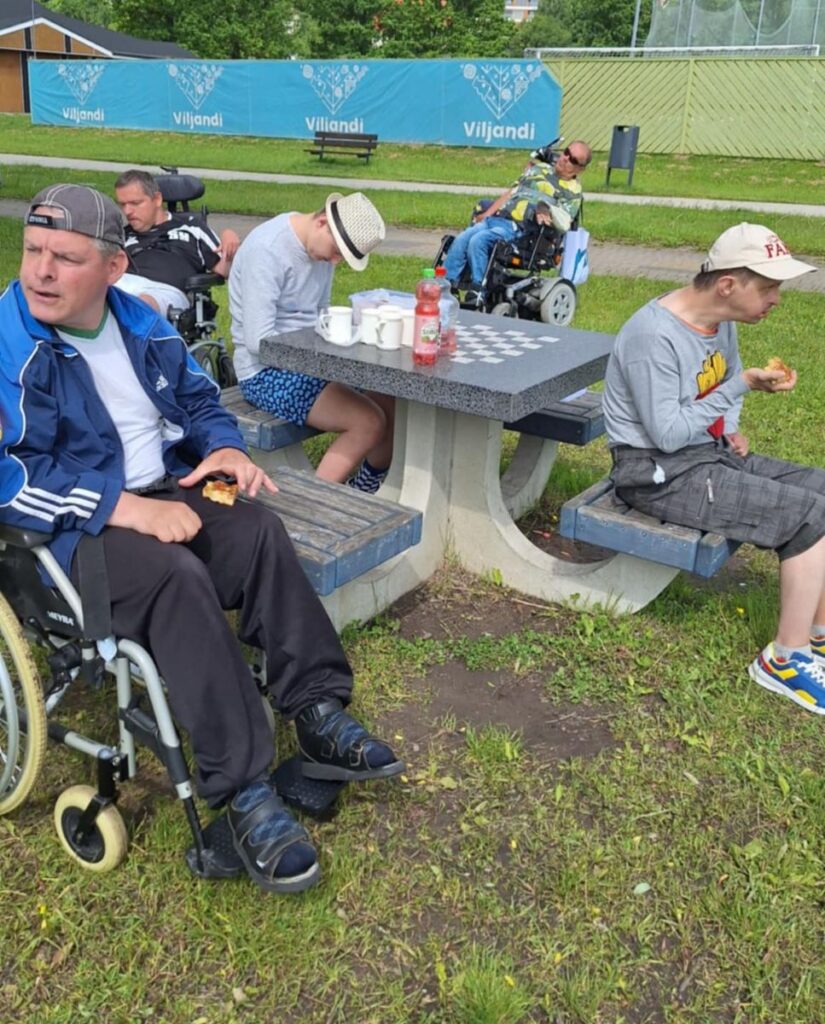 Inimesed istuvad toolidel ja ratastoolis õues laua ümber, söövad ja joovad. Taustal on Viljandi silt ja roheline muruplats.