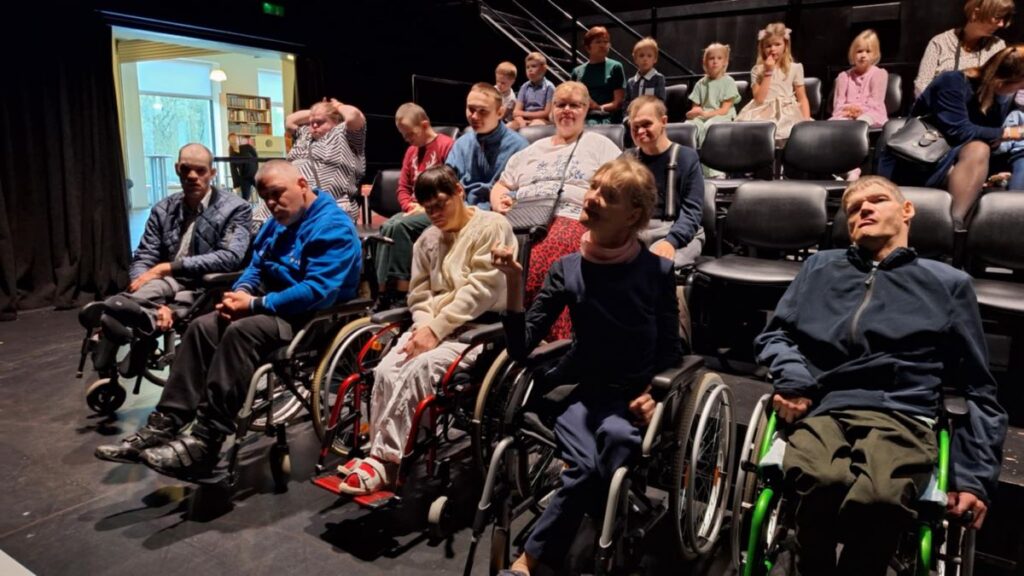 Rühm inimesi, sealhulgas ratastoolikasutajaid, istub saalis ja jälgib laval toimuvat, taga istuvad ka lapsed.