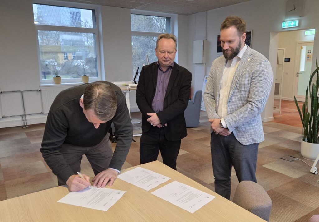 Kontoriruumis kirjutab üks mees laua taga koostöökokkuleppele alla, samal ajal kui kaks meest seisavad kõrval ja jälgivad. Laual on laiali mitu allkirjastamiseks valmis dokumenti ning taustal paistavad aknad ja istumisnurk.