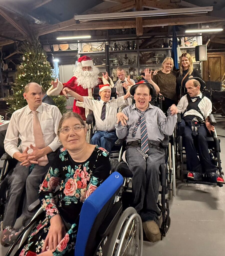 Jõulupidulik seltskond koos jõuluvana ja kuusega taustal; osalejad on rõõmsas meeleolus, mitmed ratastoolis