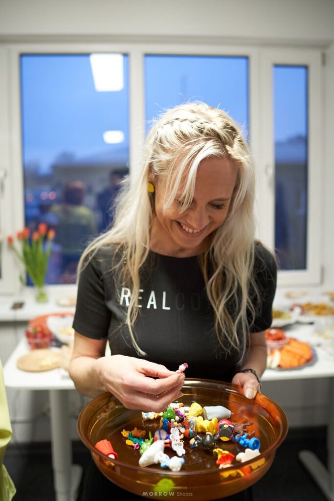 Naeratav naine vaatab kandikul olevaid värvilisi väikseid mänguasju ja esemeid heledas ruumis akna ees.