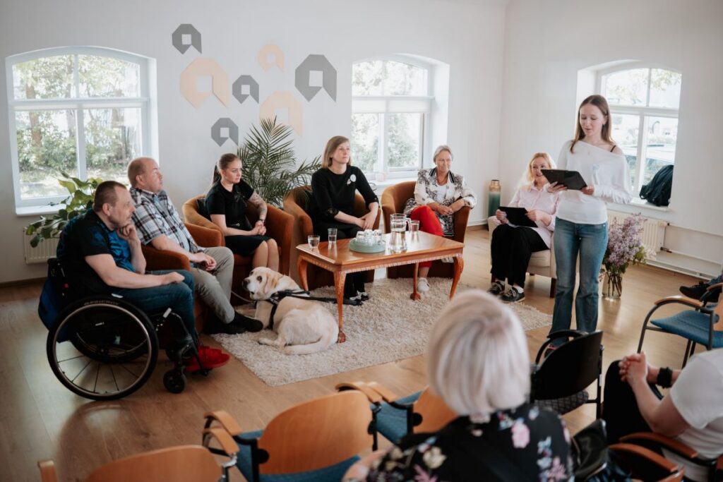Inimesed istuvad valgusküllases ruumis poolkaares, üks osaleja on ratastoolis ning põrandal lamab juhtkoer. Noorem naine seisab ja loeb tahvelarvutist arutelu mõtteid ette